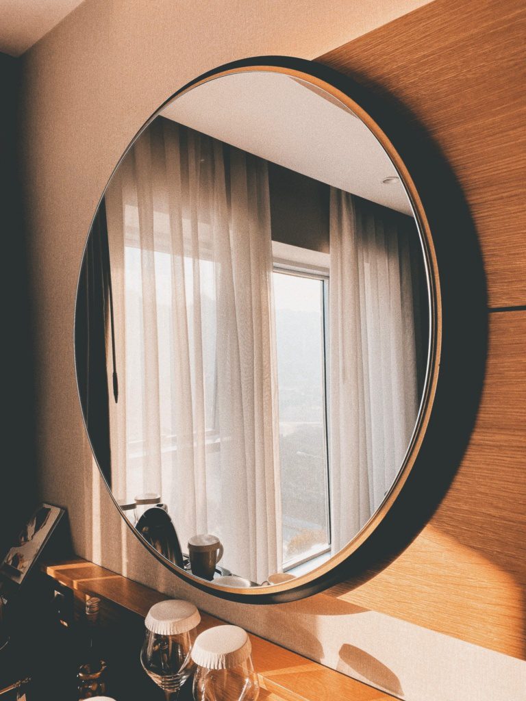 Ronde spiegel in warme hotelkamer met houten details, zachte gordijnen en zonlicht dat naar binnen valt.